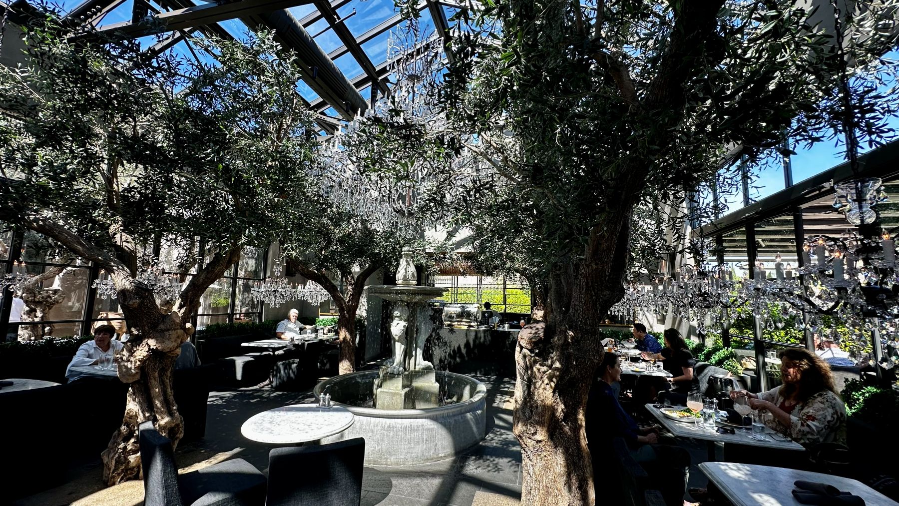 Wide interior view of RH Yountville dining room with glass architecture, large scale furnishings, crystal chandelier and olive trees creating an indoor-outdoor living room atomosphere